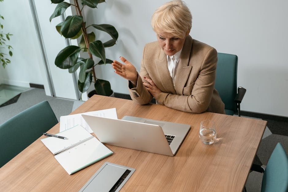 Professional woman in a blazer working on a laptop at a wooden desk in a modern office setting.