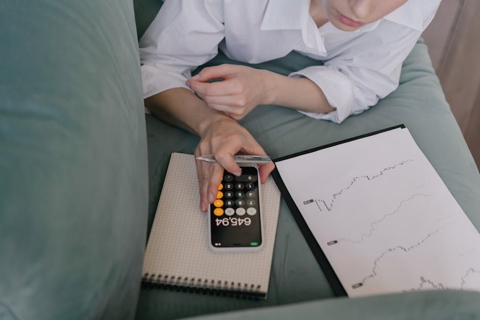 A woman works indoors using a smartphone calculator and analyzing financial graphs, symbolizing modern finance.